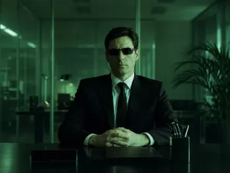 Agent Mann seated at a desk in a green-tinted office, wearing sunglasses and an earpiece.