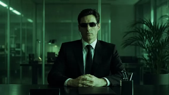 Agent Mann seated at a desk in a green-tinted office, wearing sunglasses and an earpiece.